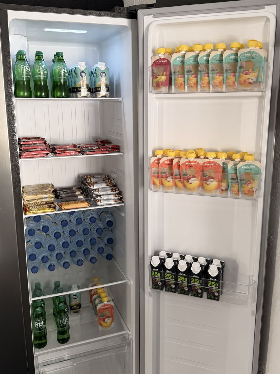Frigorífico do estúdio cheio: águas, refrigerantes, barras de proteína, papas de fruta e batidos de 25g de proteína.