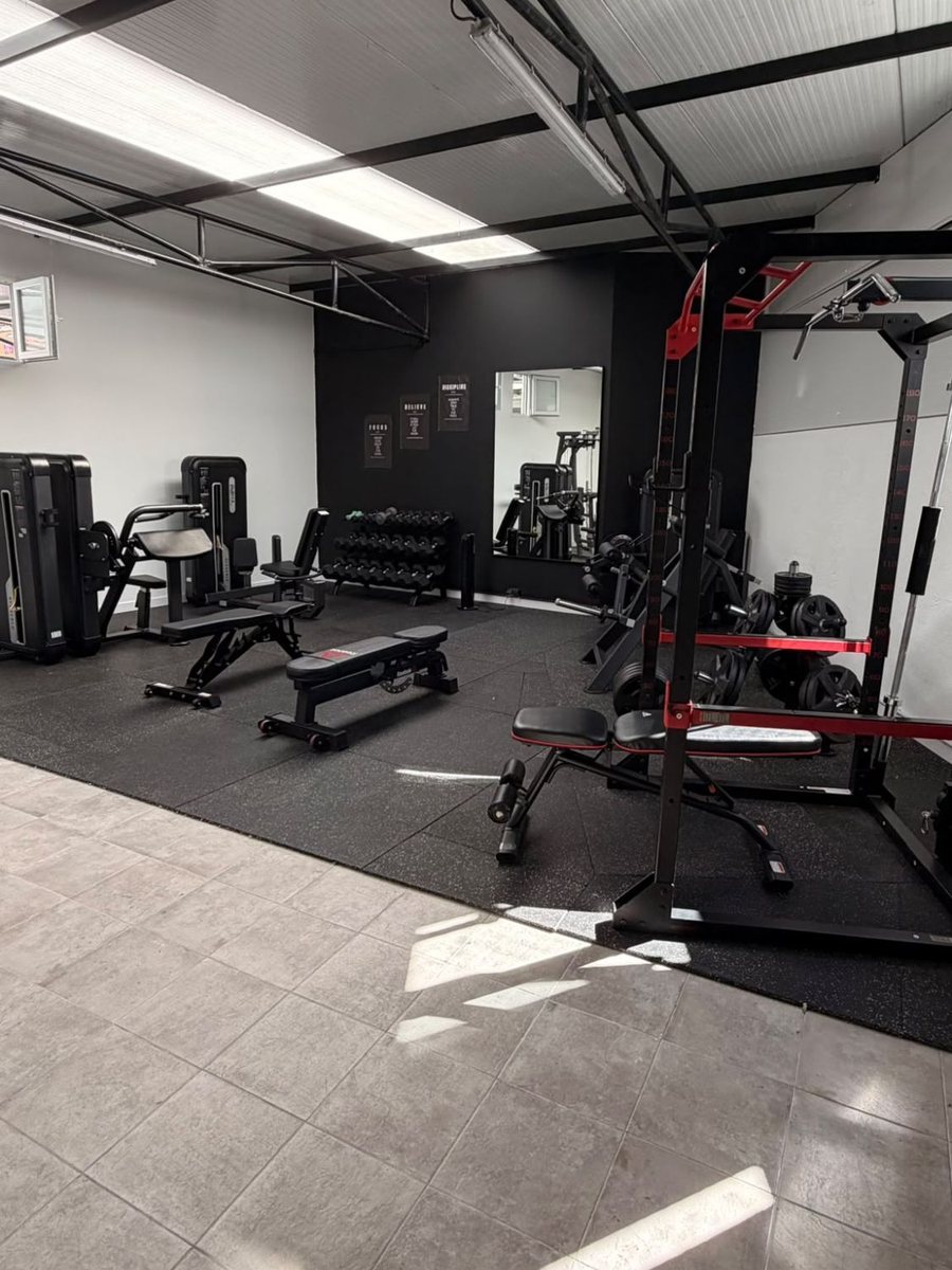 A sala de treino do estúdio: rack com barra e discos, bancos reguláveis, máquinas de cabos, halteres e parede de espelho.